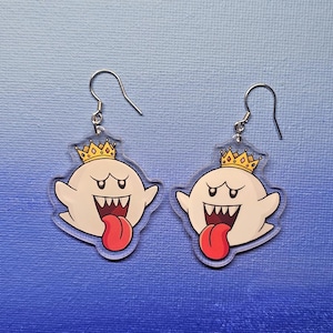 May include: A pair of ghost-shaped earrings. Each earring features a white ghost with a red tongue, a wide open mouth, and a gold crown. The earrings have silver-colored fish hook ear wires.