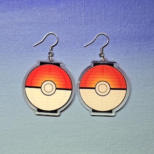 May include: A pair of acrylic earrings featuring a design reminiscent of a Poké Ball. Each earring has a red and orange gradient top, a white bottom, and a black circle in the center. The earrings hang from silver-colored hooks.