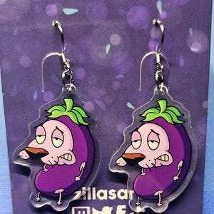 May include: A pair of clear acrylic earrings featuring a purple eggplant design with a cartoon dog face. The earrings have silver-colored hooks. The background is purple with a logo and social media icons.