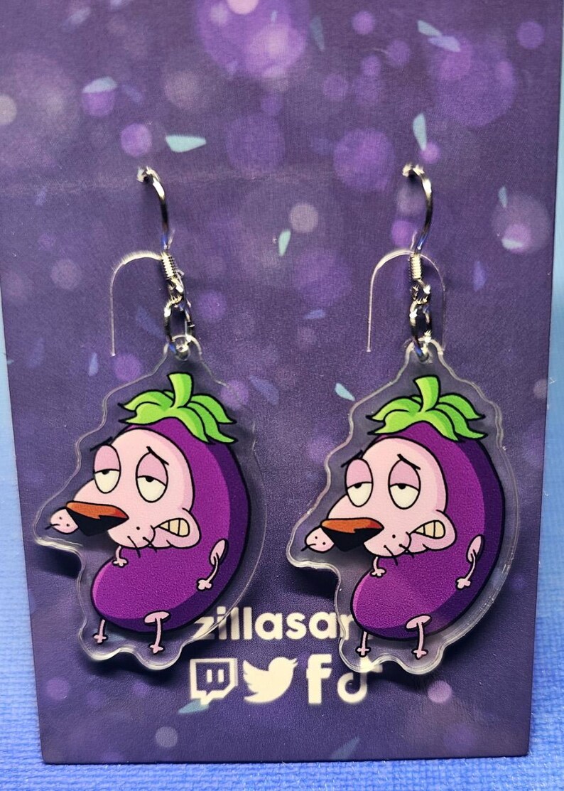 May include: A pair of clear acrylic earrings featuring a purple eggplant design with a cartoon dog face. The earrings have silver-colored hooks. The background is purple with a logo and social media icons.