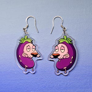 May include: A pair of clear acrylic earrings featuring a cartoon eggplant character with a sad expression. The earrings have silver-colored fish hook ear wires and a purple, green, and tan color scheme. The earrings are on a blue background.