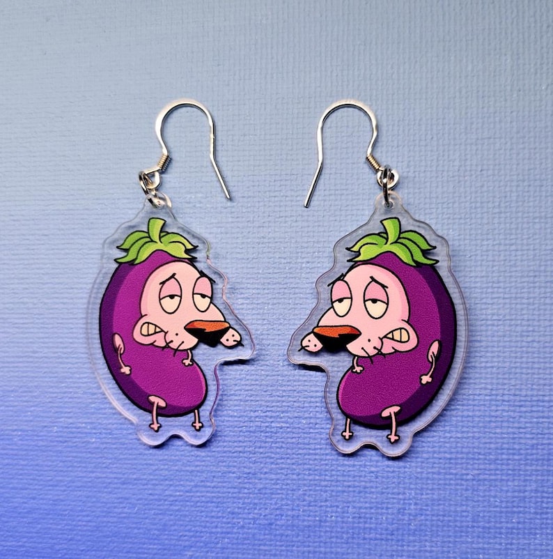 May include: A pair of clear acrylic earrings featuring a cartoon eggplant character with a sad expression. The earrings have silver-colored fish hook ear wires and a purple, green, and tan color scheme. The earrings are on a blue background.