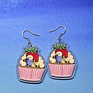 May include: A pair of clear acrylic earrings featuring a cupcake design. Each earring has a pink cupcake liner, yellow and black stripes, white frosting, a red strawberry, and a blue character.