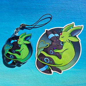 Op de afbeelding: Een groene en zwarte sleutelhanger met een cartoonfiguur met twee Pok&eacute;mon-personages, een groene Leafeon en een zwarte Umbreon, die elkaar knuffelen. De sleutelhanger is gemaakt van duurzaam materiaal en heeft een zwart koord.