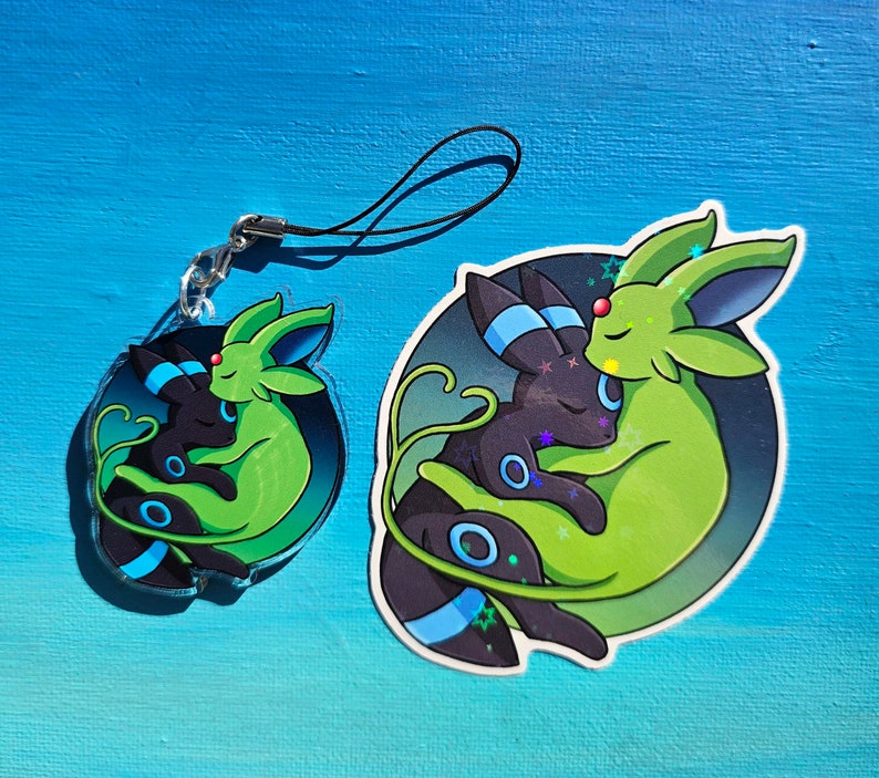 Op de afbeelding: Een groene en zwarte sleutelhanger met een cartoonfiguur met twee Pok&eacute;mon-personages, een groene Leafeon en een zwarte Umbreon, die elkaar knuffelen. De sleutelhanger is gemaakt van duurzaam materiaal en heeft een zwart koord.