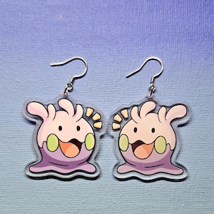 May include: A pair of clear acrylic earrings featuring a cartoon character with a purple body, light pink head, and green cheeks. The character has a smiling face with an open mouth and yellow accents. The earrings have silver-colored fish hook closures.