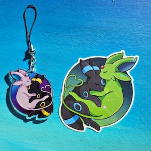 Op de afbeelding: Een zwarte en groene anime-personage hanger met een roze en zwarte anime-personage hanger op een blauwe achtergrond. De hangers zijn bevestigd aan een zwart touwtje.