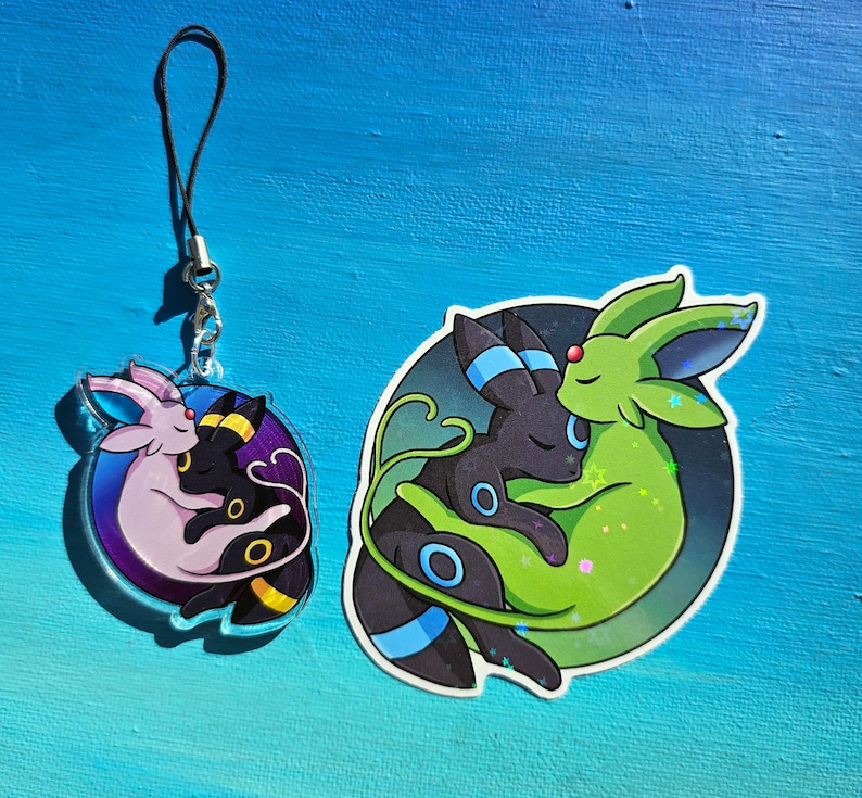 Op de afbeelding: Een zwarte en groene anime-personage hanger met een roze en zwarte anime-personage hanger op een blauwe achtergrond. De hangers zijn bevestigd aan een zwart touwtje.