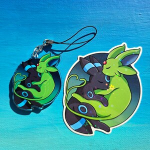 Op de afbeelding: Een sleutelhanger met een zwart en groen cartoonfiguur, dat wordt omhelsd door een zwart en blauw figuur. De sleutelhanger is op een blauwe achtergrond.