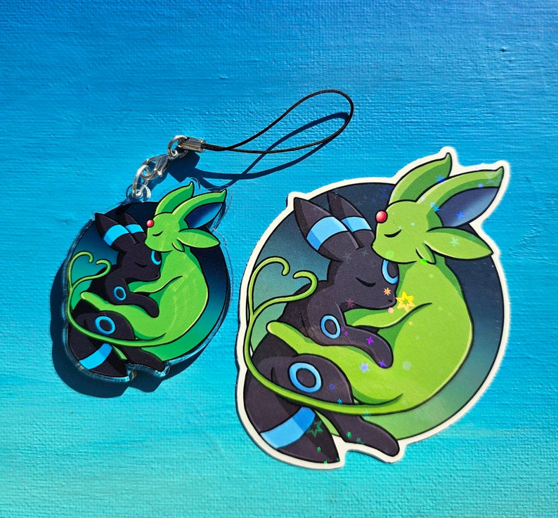 Op de afbeelding: Een sleutelhanger met een zwart en groen cartoonfiguur, dat wordt omhelsd door een zwart en blauw figuur. De sleutelhanger is op een blauwe achtergrond.