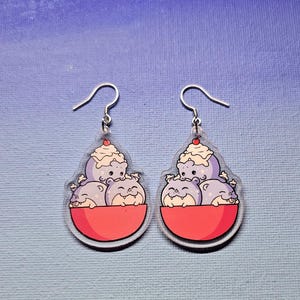 May include: A pair of dangle earrings featuring a cartoon design. Each earring has a red bowl with three purple creatures and a scoop of ice cream with a cherry on top. The earrings have silver-colored fish hook closures.