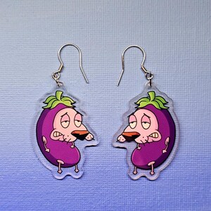 May include: A pair of dangle earrings featuring a cartoon character design. Each earring is shaped like a purple eggplant with a green leafy top and a cartoon dog face. The earrings have silver-colored fish hook ear wires.