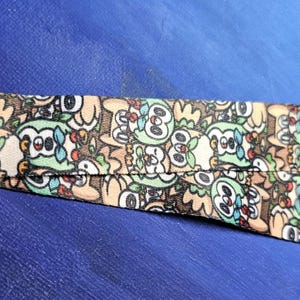May include: A lanyard with a repeating pattern of cartoon owls in shades of brown, green, and white. The lanyard has a metal clasp and a split strap design. The background is a solid blue.