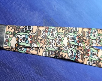 May include: A lanyard with a repeating pattern of cartoon owls in shades of brown, green, and white. The lanyard has a metal clasp and a split strap design. The background is a solid blue.