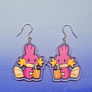 May include: A pair of clear acrylic earrings featuring a pink cartoon character with yellow accents, drinking from a juice cup. The earrings have silver-colored fish hook ear wires.
