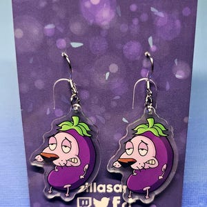 May include: Acrylic earrings featuring a cartoon eggplant character with a worried expression. The earrings are purple with green accents and silver hooks. The text "acrylic earrings" is at the top.
