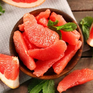 May include: A wooden bowl filled with juicy, red grapefruit segments, garnished with fresh mint leaves. Slices of grapefruit are arranged around the bowl on a wooden surface, creating a vibrant and appetizing display.