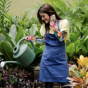 Può includere: Una donna con un grembiule blu e una camicia gialla innaffia le piante con un annaffiatoio verde. Le sue braccia sono decorate con tatuaggi floreali colorati. La scena è ambientata in un lussureggiante giardino con varie piante e fogliame verde.