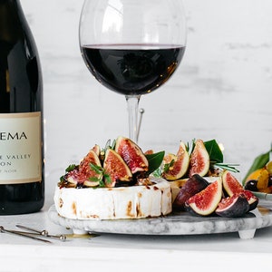 May include: A bottle of La Crema wine next to a wine glass filled with red wine. A marble plate holds a wheel of brie topped with figs and herbs. Olives and lemons are also visible, suggesting a wine and cheese pairing.