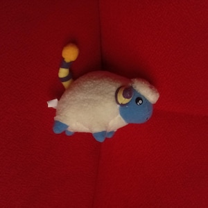 May include: A plush toy shaped like a creature with a white fuzzy body, a blue face, and blue feet. It features a yellow and blue striped tail with a yellow pom-pom at the end, and a small white tag protruding from the side of its rounded body.