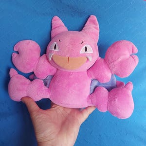 Peut inclure: Une peluche rose avec de grandes griffes, des oreilles et un visage souriant. Le jouet a des accents blancs sur les yeux et les dents, et une bouche orange clair. La peluche est tenue dans une main sur un fond bleu.