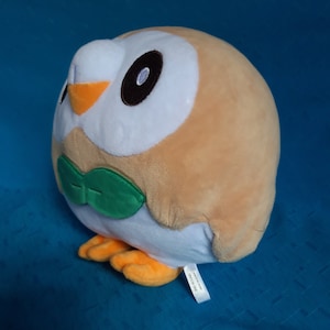 May include: A soft owl toy in tan and white, with large black eyes and an orange beak. It has green leaf-shaped details and orange feet. The plush toy has a rounded body. A small tag is attached to the owl.