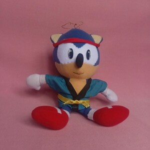 以下が含まれることがあります： 空手着を着たソニック・ザ・ヘッジホッグのぬいぐるみ。青いハリネズミは赤いヘアバンド、白い手袋、赤い靴をしています。空手着は、黒い襟と黄色のベルトが付いたティール色です。ピンク色の背景にあります。