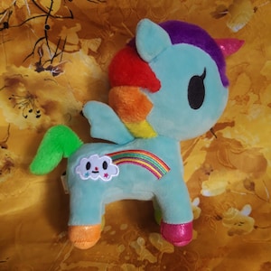 Könnte beinhalten: Ein hellblaues Einhorn-Plüschtier mit Regenbogenmähne und -schwanz. Auf der Seite befindet sich ein Regenbogen- und Wolkenmotiv. Das Einhorn hat pinke Hufe, ein lilafarbenes Horn und große schwarze Augen.