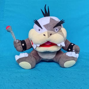 May include: A plush toy character with a white and grey head, yellow snout, and brown body. The character has black spikes, white teeth, and holds a grey club with a red ball. The toy is on a blue background.
