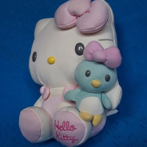 Peut inclure: Un jouet en peluche Hello Kitty avec un nœud rose et un petit jouet en peluche de pingouin bleu. Le jouet Hello Kitty porte une robe rose et a les mots "Hello Kitty" brodés sur sa jambe.
