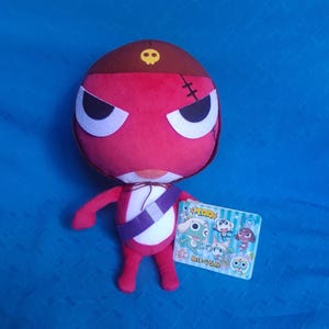 May include: A red and white plush toy wearing a brown pirate hat with a skull and crossbones design. The toy has a purple sash and is holding a card with Japanese text and images of other plush toys.