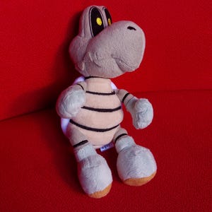 Könnte beinhalten: Ein Dry Bones Plüschtier aus der Super Mario Videospielserie. Das Spielzeug hat einen hellgrauen Kopf und Gliedmaßen, einen beigen Körper mit schwarzen Streifen und gelben Augen. Das Spielzeug befindet sich auf einer roten Oberfläche.