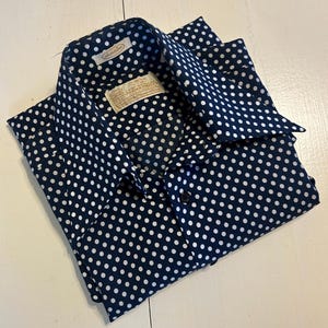 Puede incluir: Una camisa de manga corta azul marino con estampado de lunares blancos. La camisa tiene cuello abotonado y cierre de botón.
