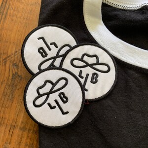 以下が含まれることがあります： 黒いTシャツに白いリブが付いた、黒い輪郭と「LB」の文字、そしてスタイリッシュなカウボーイハットのデザインが施された白い刺繍パッチが3つあります。