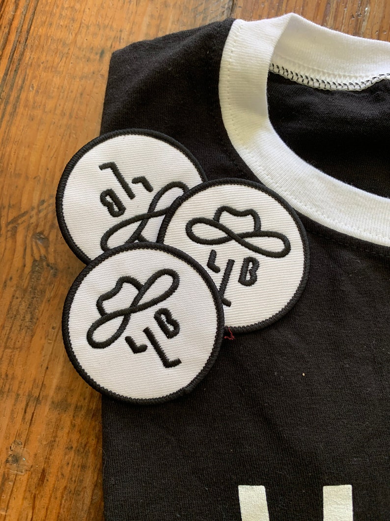 以下が含まれることがあります： 黒いTシャツに白いリブが付いた、黒い輪郭と「LB」の文字、そしてスタイリッシュなカウボーイハットのデザインが施された白い刺繍パッチが3つあります。