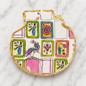 Puede incluir: Un posavasos de cer&aacute;mica blanca con forma org&aacute;nica irregular y borde pintado en oro met&aacute;lico. La superficie presenta una cuadr&iacute;cula de ilustraciones dibujadas a mano que incluyen un p&aacute;jaro morado, jarrones florales y los n&uacute;meros 2 y 3.