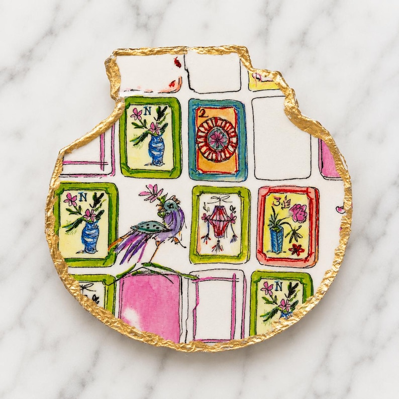 Puede incluir: Un posavasos de cer&aacute;mica blanca con forma org&aacute;nica irregular y borde pintado en oro met&aacute;lico. La superficie presenta una cuadr&iacute;cula de ilustraciones dibujadas a mano que incluyen un p&aacute;jaro morado, jarrones florales y los n&uacute;meros 2 y 3.