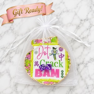 Puede incluir: Una bolsa de organza blanca con cord&oacute;n que contiene una concha de ostra decorativa con borde dorado. La concha presenta un gr&aacute;fico colorido con bamb&uacute;, un p&aacute;jaro y el texto Dot Crack BAM, coronado por dos peque&ntilde;os dados morados con puntos blancos.