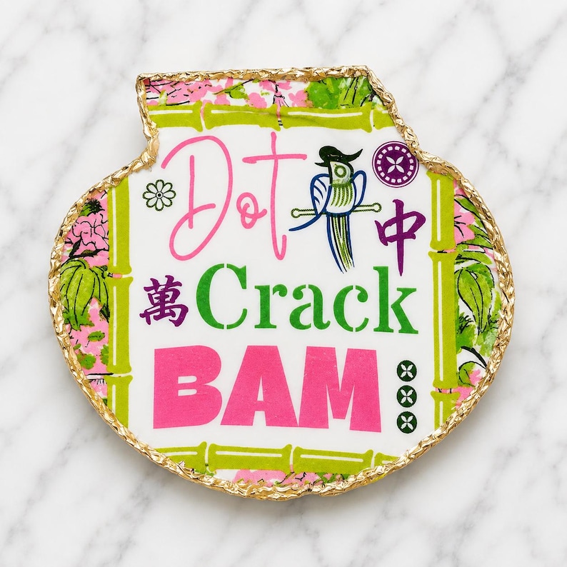 Puede incluir: Una bandeja decorativa con forma de concha y borde pintado en oro. El centro presenta el texto Dot, Crack y BAM en rosa y verde, junto a una ilustraci&oacute;n de un p&aacute;jaro azul, tallos de bamb&uacute; verdes, motivos florales rosas y caracteres chinos.