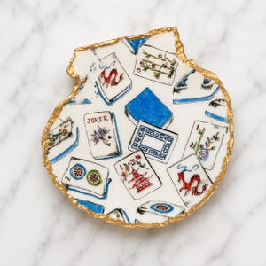 Puede incluir: Un plato decorativo blanco con forma de concha y borde irregular pintado en dorado. La superficie presenta un patr&oacute;n repetitivo de fichas de Mahjong coloridas, incluyendo un comod&iacute;n, dragones rojos, bamb&uacute; y pagodas en un estilo pintado a mano.
