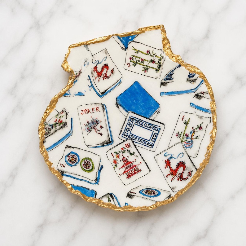 Puede incluir: Un plato decorativo blanco con forma de concha y borde irregular pintado en dorado. La superficie presenta un patr&oacute;n repetitivo de fichas de Mahjong coloridas, incluyendo un comod&iacute;n, dragones rojos, bamb&uacute; y pagodas en un estilo pintado a mano.