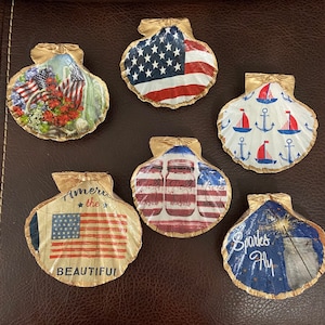 May include: A collection of six decorative seashells with gold-colored edges. The shells feature patriotic designs, including the American flag, sailboats, anchors, and the text "America the Beautiful" and "Sparks Fly".