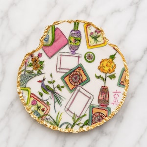 Puede incluir: Un posavasos circular de cer&aacute;mica blanca con un borde irregular de pan de oro. La superficie presenta un patr&oacute;n pintado a mano de varios marcos rectangulares, jarrones con flores, un p&aacute;jaro y peque&ntilde;os motivos circulares, incluyendo caracteres chinos en rojo.