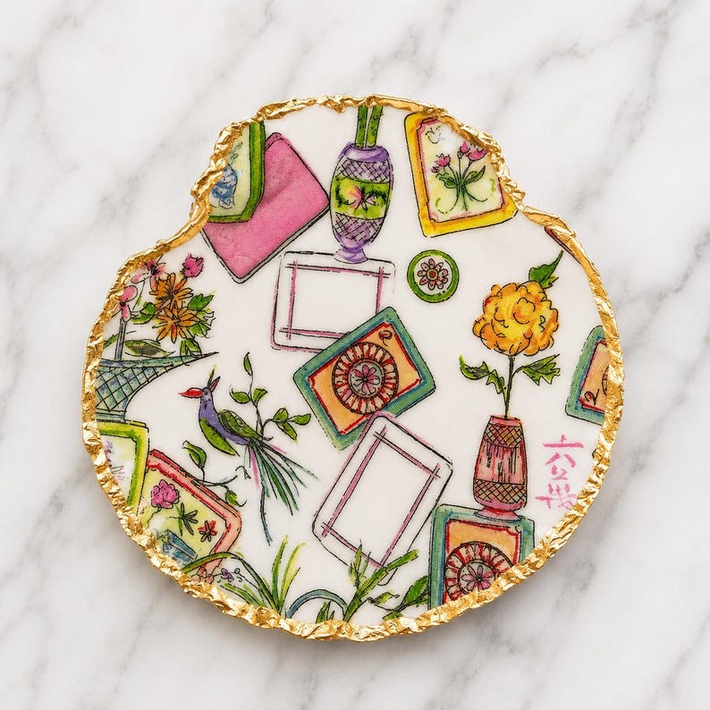 Puede incluir: Un posavasos circular de cer&aacute;mica blanca con un borde irregular de pan de oro. La superficie presenta un patr&oacute;n pintado a mano de varios marcos rectangulares, jarrones con flores, un p&aacute;jaro y peque&ntilde;os motivos circulares, incluyendo caracteres chinos en rojo.