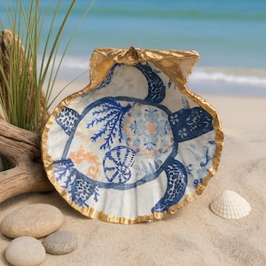 May include: A decoupage Lions Paw seashell with a gold-painted rim. The interior features a blue and white floral and turtle design. The shell rests on sand with small stones and a piece of driftwood, with the ocean in the background.