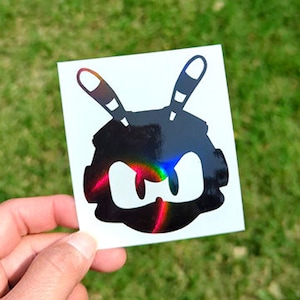 Puede incluir: Una pegatina en blanco y negro de una cabeza de robot con antenas. El robot tiene ojos grandes y enojados y un cuerpo negro redondeado.