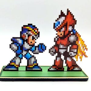 May include: A pixel art figurine set featuring two characters from the Mega Man video game series. The character on the left is a blue and white robot with a red helmet and a blue and white arm. The character on the right is a red and white robot with a red helmet and a yellow flame effect. The figurines are standing on a green base.