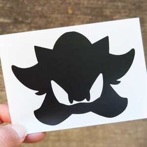 May include: Black silhouette of a cartoon character with sharp teeth and a spiky head. The character is facing forward with its mouth open.