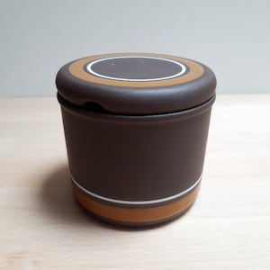 Könnte beinhalten: Eine braune Zuckerdose aus Keramik mit Deckel. Der Deckel hat ein weißes und orangefarbenes Kreisdesign. Die Schüssel hat einen weißen und orangefarbenen Streifen um den Boden.