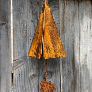 May include: A rustic, metal wind chime with a triangular top and a leaf-shaped bottom. The chime has a weathered, orange-brown finish, and hangs from a chain. The background is a weathered wooden fence.